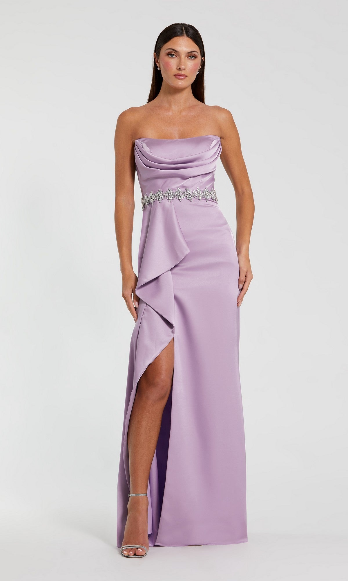 A woman in the lilac Mac Duggal 44133 long formal dress, featuring a strapless satin design with an embellished belt, draped front, thigh-high slit, and silver heels stands against a plain background.